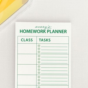 May include: A white homework planner with green text that says "avery's HOMEWORK PLANNER" and has the headings "CLASS" and "TASKS". The planner has 10 rows for listing classes and tasks. There are 7 colourful clothes pegs on a white surface.