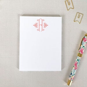 May include: A white notepad with a pink letter H monogram on the front. The notepad is on a light beige surface with a gold pen and two gold paper clips.