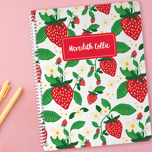 May include: A spiral-bound notebook with a white background covered in a pattern of red strawberries, green leaves, and white flowers. The notebook has a red rectangular label with the text "Meredith Collie". Three pens in shades of pink and yellow are on a pink surface.
