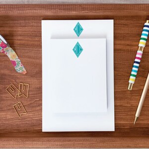 May include: A wooden tray with a stack of white notepad paper with a turquoise diamond monogram on the top sheet. The tray also contains a colorful striped pen, a wooden pencil, and a roll of floral patterned washi tape.