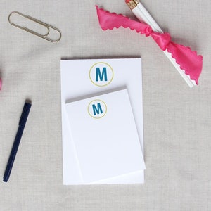 May include: Two white notepad sets with a blue letter "M" in a gold circle on the front. The notepad sets are on a light gray fabric surface with a pink ribbon, two pencils, a gold paperclip, and pink tulips.