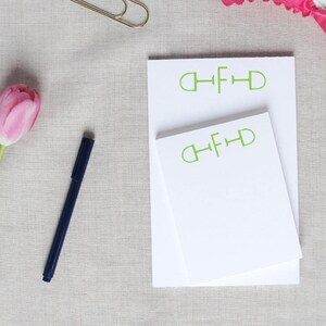 May include: Two white notepad sets with green "CHFD" lettering on a light gray fabric background. A pink flower, a blue pen, and a gold paper clip are also visible.