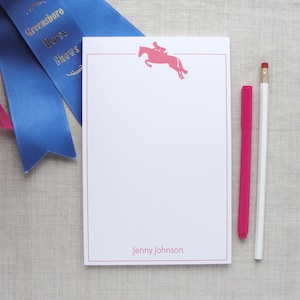 May include: A white notepad with a pink horse and rider silhouette, a red border, and the text "Jenny Johnson" at the bottom. Two pens, one white and one pink, are next to the notepad.