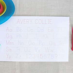 May include: A white sheet of paper with dotted lines for tracing the alphabet and numbers. The letters are in different colors. The paper has the name Avery Collie written at the top.