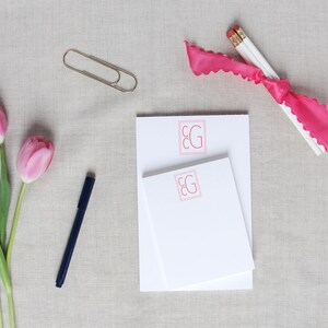 May include: Two white notepads with pink and red "EG" monogrammed on the front. The notepads are on a light gray fabric surface with a pink ribbon, two pencils, a gold paperclip, and pink tulips.