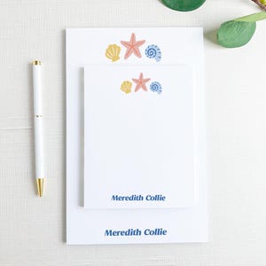 May include: White stationery set featuring seashell and starfish illustrations in yellow, pink, and blue. The set includes a notepad and a larger sheet, with the name "Meredith Collie" printed in blue.