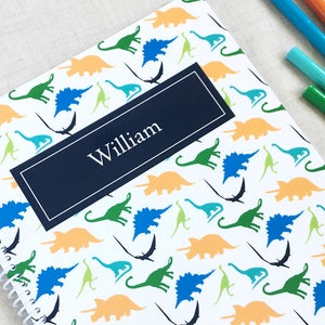 May include: A white spiral notebook with a blue and orange dinosaur pattern. The notebook has a navy blue label with the name "William" printed in white.