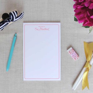 May include: A white notepad with a red border and the text "Thank you for the gift of" and "Erin Blanchard" in pink cursive. The notepad is on a beige fabric surface with a blue pen, a black and white striped box, a pink eraser, and a bundle of pencils tied with a gold ribbon.