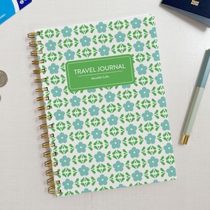 Peut inclure: Un carnet de voyage à reliure spirale avec une couverture blanche ornée d'un motif répétitif de fleurs bleu clair et de feuilles vertes. Une étiquette rectangulaire verte au centre affiche le texte TRAVEL JOURNAL et Meredith Collie en blanc.