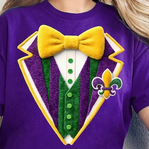 May include: Purple t-shirt featuring a graphic of a tuxedo jacket with a yellow bow tie, green and purple striped vest, and a fleur-de-lis. The design has a textured, 3D appearance, perfect for Mardi Gras celebrations.