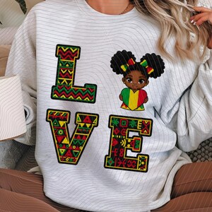 Puede incluir: Una sudadera gris claro con la palabra "LOVE" en un diseño colorido y estampado. La "O" se reemplaza con una ilustración de una niña sosteniendo un corazón. El diseño incorpora colores rojo, amarillo, verde y negro.