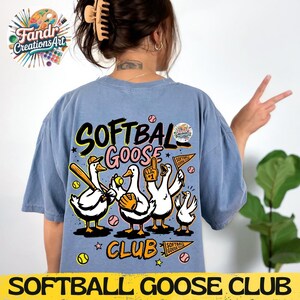 May include: A light blue t-shirt featuring a cartoon design of geese playing softball. The text "SOFTBAL GOOSE CLUB" is displayed in a playful font. The geese are depicted with baseball bats, gloves, and a pennant.
