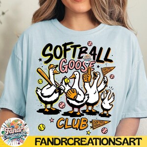 May include: Light blue t-shirt with a graphic of cartoon geese playing softball. The design includes the words "Softball Goose Club" in black and yellow, with baseballs and stars. The brand name "Fandr Creations Art" is at the bottom.