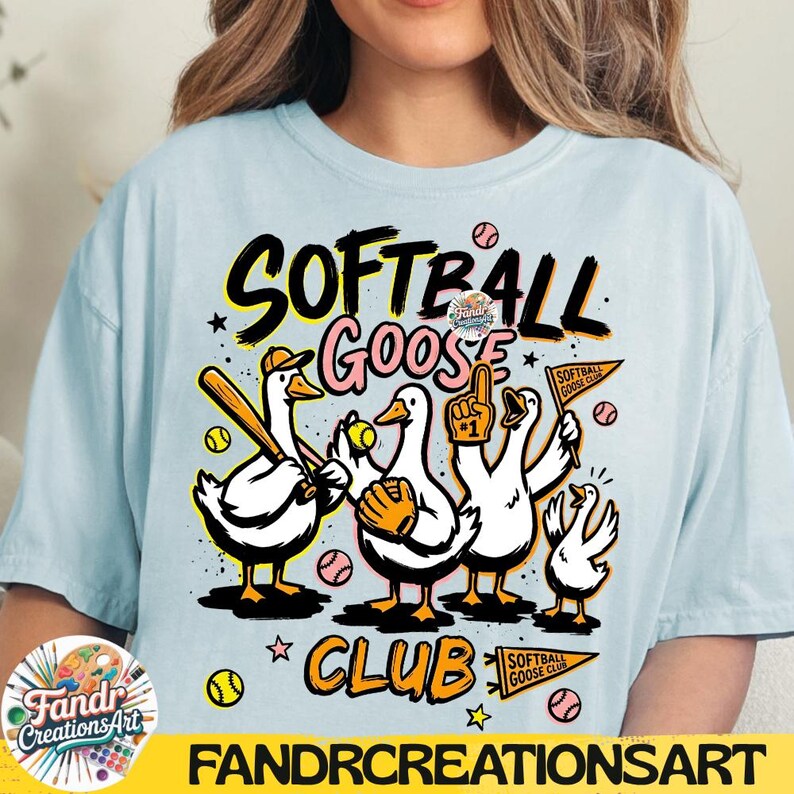 May include: Light blue t-shirt with a graphic of cartoon geese playing softball. The design includes the words "Softball Goose Club" in black and yellow, with baseballs and stars. The brand name "Fandr Creations Art" is at the bottom.
