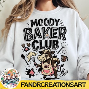 May include: White sweatshirt with the text "MOODY BAKER CLUB" in black. A cartoon cow wearing a pink apron and holding a whisk is featured. Baking-themed graphics like a spoon, measuring cup, and splatters are also included.