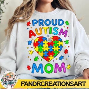 Op de afbeelding: Witte sweatshirt met de woorden "PROUD AUTISM MOM" in kleurrijke letters. Een hartvormig puzzelstukontwerp in het midden heeft verschillende kleuren. De sweatshirt heeft ook de tekst "FANDRCREATIONSART" onderaan.