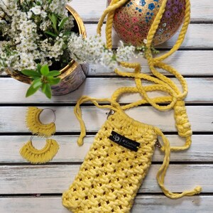 Può includere: Una borsa per telefono in macramè giallo con cinturino abbinato e un paio di orecchini con frange. La borsa ha una piccola etichetta nera con la scritta "d'Crafted". Una palla decorativa dipinta e un vaso di fiori bianchi sono anche nell'immagine.