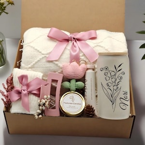 Könnte beinhalten: Eine Geschenkbox gefüllt mit einer cremefarbenen Decke mit rosa Schleife, einem weißen Becher mit floralem Design und dem Namen "Nori", einer Kerze und anderen kleinen Geschenken. Die Box ist mit braunem Karton ausgekleidet.