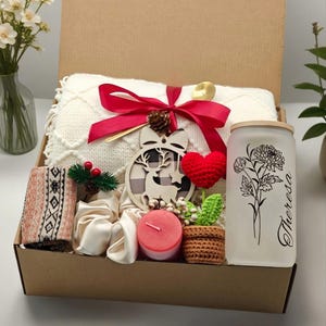 May include: A gift box filled with a cream-colored blanket, red ribbon, and decorative items. Includes a wooden ornament, a red heart, a candle, a glass with floral design, and a pair of socks. The box is brown.
