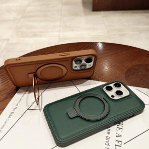 Op de afbeelding: Twee telefoonhoesjes met ringhouders. Eén is bruin, de andere is donkergroen. Beide hoesjes hebben een gestikte rand en een cirkelvormige ringhouder. De bruine hoes heeft een goudkleurige ring die als standaard kan worden gebruikt.