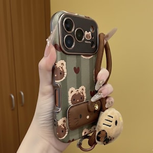 May include: A green and brown phone case featuring a Hello Kitty design with a matching wrist strap and a small plush charm. The case has a leather accent and a metal frame around the camera.