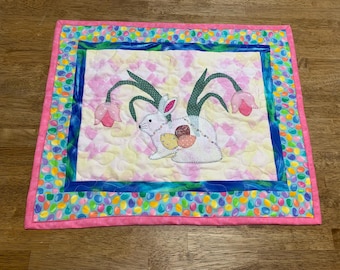 May include: A white quilted wall hanging with a pink border features a white bunny with green leaves and pink flowers. The border is decorated with colorful jelly bean shapes.