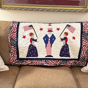 May include: A patriotic pillow featuring a quilted design with Uncle Sam and two figures holding American flags. The pillow has a white center with red stars and a blue border, and is surrounded by a red, white, and blue flag pattern.