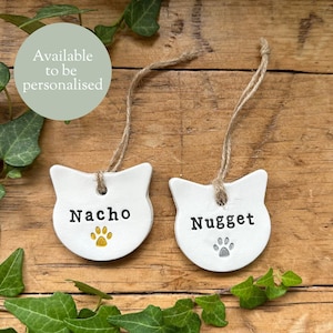May include: Two white ceramic cat-shaped ornaments with jute twine hangers. One says "Nacho" with a gold paw print, the other says "Nugget" with a grey paw print. The text "Available to be personalised" is in a green circle.