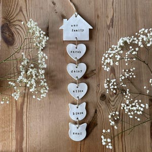 May include: White ceramic home decor hanging with the words "our family" on a house shape, followed by four heart shapes with the names "sarah", "daisy", "alice", and two cat shapes with the names "kiara" and "zemi" written on them.