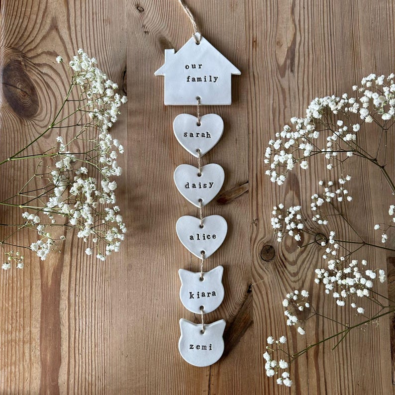 May include: White ceramic home decor hanging with the words "our family" on a house shape, followed by four heart shapes with the names "sarah", "daisy", "alice", and two cat shapes with the names "kiara" and "zemi" written on them.