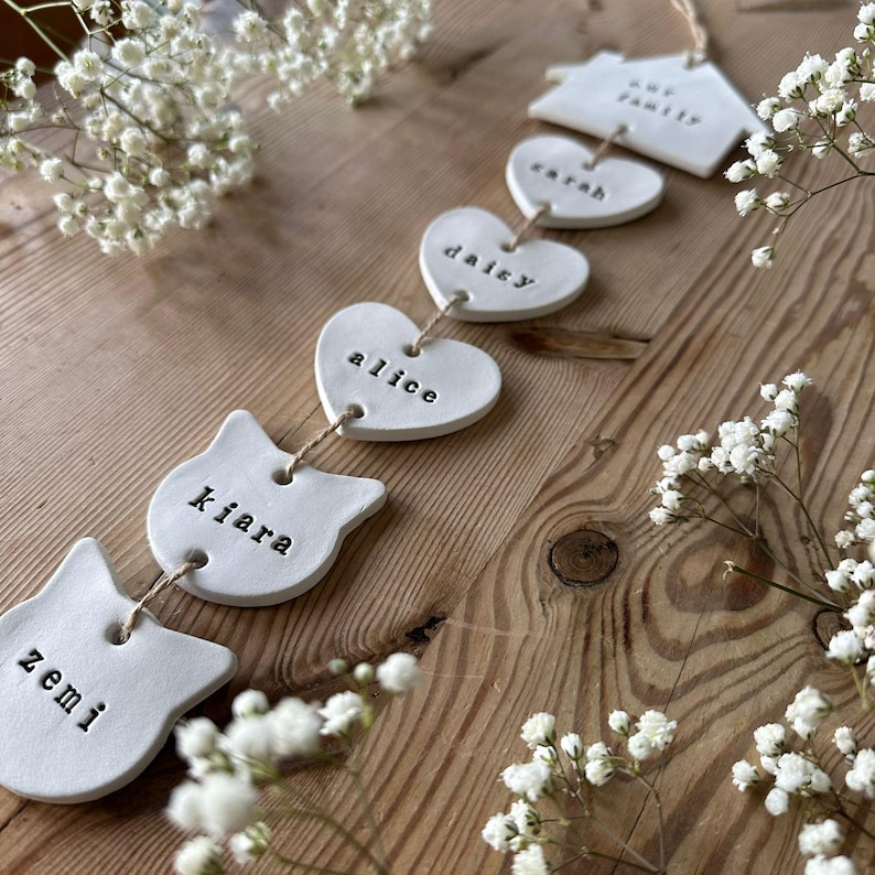 May include: A white ceramic garland with the words "Our Family", "Sarah", "Daisy", "Alice", "Kiara", and "Zemi" written on each piece. The garland is strung together with twine and is hanging on a wooden surface.