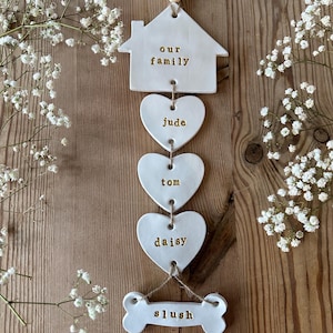 May include: White ceramic house shaped ornament with the text "our family" hanging from a string with white ceramic hearts with the names "jude", "tom", and "daisy" and a white ceramic bone with the name "slush" hanging from it.