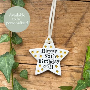 May include: A white star-shaped ornament with gold star detailing and the words "Happy 70th Birthday Gill". It hangs from a cream ribbon. The background is a wooden surface with green leaves.