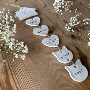 May include: A white ceramic garland with the words "our family", "sarah", "dalcy", "alice", "kiara", and "zemi" written on each piece. The garland is strung together with twine.