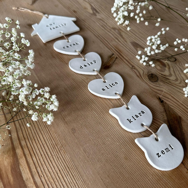 May include: A white ceramic garland with the words "our family", "sarah", "dalcy", "alice", "kiara", and "zemi" written on each piece. The garland is strung together with twine.