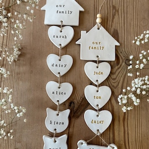 May include: White ceramic ornaments with the words "our family" on two houses, and names like "Sarah", "Daisy", "Alice", "Kiara", "Zemi", "Jude", "Tom", "Daisy", and "Slush" on hearts, a cat, and a bone.