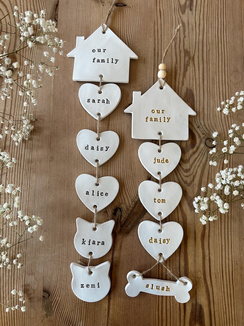 May include: White ceramic ornaments with the words "our family" on two houses, and names like "Sarah", "Daisy", "Alice", "Kiara", "Zemi", "Jude", "Tom", "Daisy", and "Slush" on hearts, a cat, and a bone.
