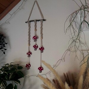 Puede incluir: Un tapiz de macramé de pared con forma triangular y tres hebras de cuerda anudada. Cada hebra tiene cuentas de madera y adornos con forma de hongo rojo y blanco.