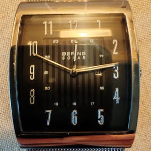 May include: A silver watch with a black face and silver hands. The watch has a rectangular face with a black and white striped pattern. The watch face has the numbers 1 through 12 and the words "BEFING SOLAR".