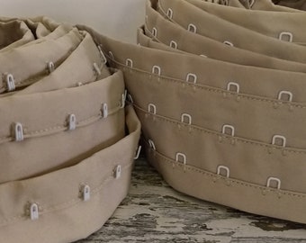 Könnte beinhalten: Zwei Rollen beigefarbenes BH-Band mit Haken und Ösen. Das Band hat weiße Kunststoffhaken und Ösen.