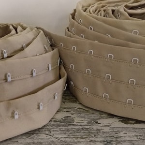 Può includere: Due rotoli di nastro per reggiseno beige con ganci e occhielli. Il nastro è dotato di ganci e occhielli in plastica bianca.