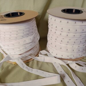 Peut inclure: Deux bobines de tissu blanc avec de petits trous espacés uniformément. Le tissu est enroulé autour de bobines en carton avec des étiquettes.