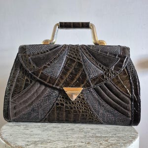 May include: A structured handbag featuring a dark brown and charcoal grey patchwork design with a mix of embossed crocodile and lizard textures. It includes a metallic gold-tone top handle and a triangular gold-tone metal clasp closure.