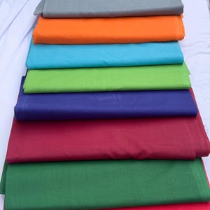 Può includere: Una pila di tessuti piegati in vari colori, tra cui giallo, verde acqua, grigio, arancione, turchese, verde, blu navy, bordeaux, verde foresta e rosso. I tessuti sono ordinatamente disposti, mostrando uno spettro di tonalità.