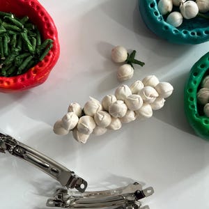 May include: A white flower garland made of small, round, white flowers. The garland is on a white surface with three silver hair clips.