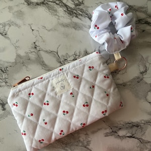 Op de afbeelding: Witte gewatteerde etui- en scrunchie-set met een kersenpatroon. De etui heeft een gouden rits en een klein label met tekst. De scrunchie en etui zijn wit met een herhalend patroon van rode kersen en groene bladeren.