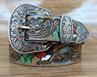 May include: A brown leather belt with a decorative copper-colored buckle and belt loop. The belt features carved floral and cactus designs, along with a painted rooster. The buckle is approximately 3 inches wide.