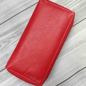 May include: A bright red leather wallet with a zipper closure. The wallet is rectangular and features the embossed name "AMELIA" on the front. The wallet is made of a textured leather material.