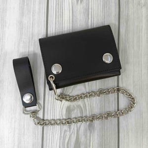 May include: Black leather wallet with a silver chain and snap closure. The wallet has a matching black leather strap with a silver snap. The chain is attached to the wallet with a silver ring.