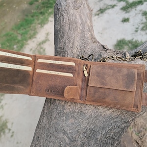 May include: Open brown leather wallet with multiple card slots, a zippered pocket, and a snap closure. The wallet has a chain attached and is displayed open, showcasing its interior compartments. The wallet is made of distressed leather.